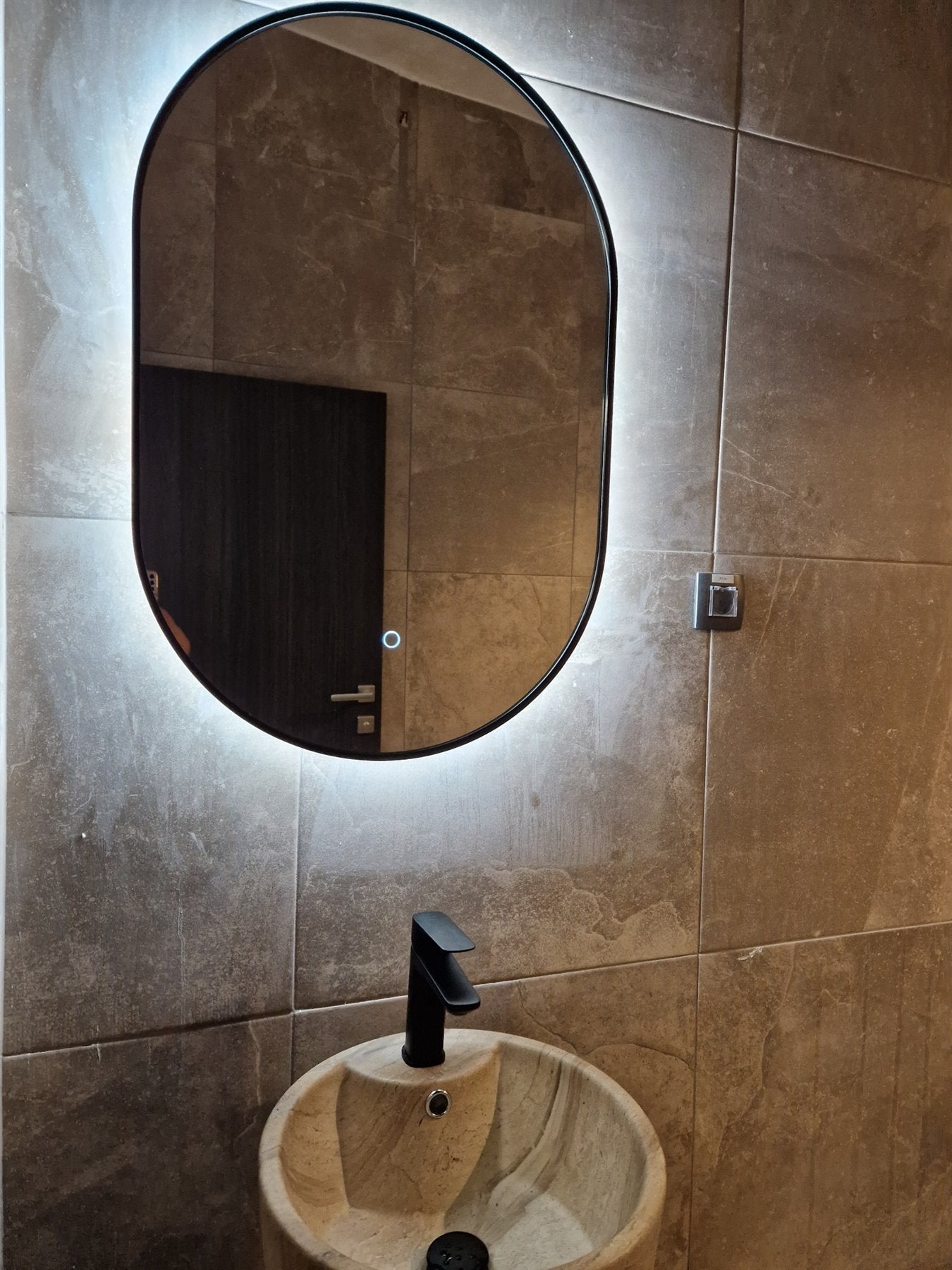 Oval mirror with a black frame and lighting above a stone washbasin.